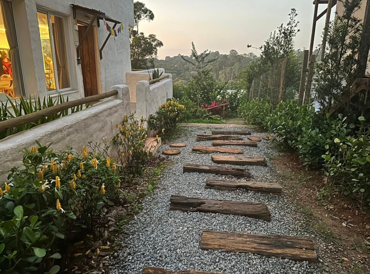 A stone pathway lined with wooden planks meanders through a garden. Flowering shrubs and yellow blooms create a colorful border, while soft gravel provides texture underfoot. A glimpse of greenery and trees can be seen in the background, enhancing the serene outdoor setting.
