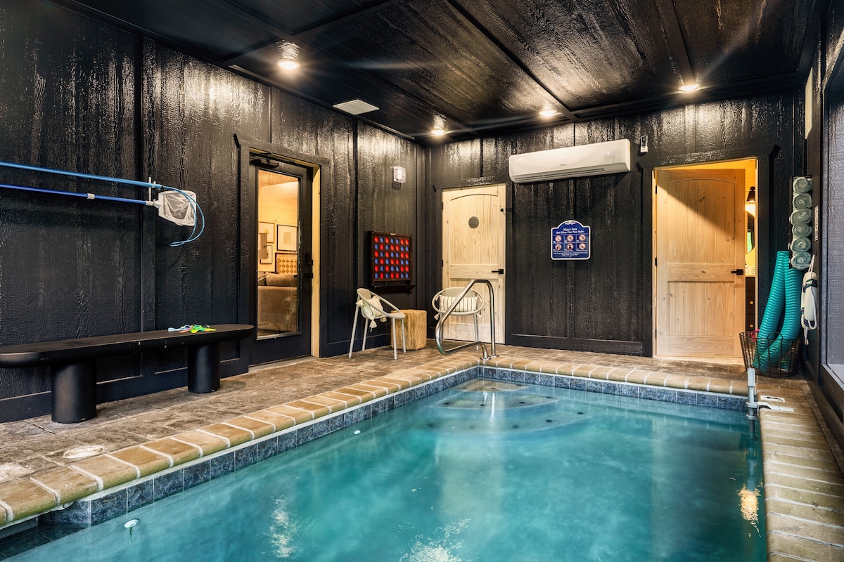 An indoor pool area features a serene swimming space with clear water. The surrounding walls are painted in a dark hue, contrasting with the light-colored doors and tiled flooring. Two lounge chairs are positioned beside the pool, while a wall-mounted drying rack adds practicality.