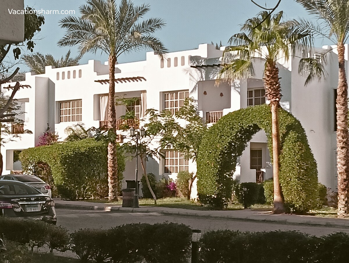 A white building with multiple balconies is surrounded by lush greenery and palm trees. Arched hedges enhance the landscape, while a parked car is visible in the foreground. The scene conveys a serene atmosphere in a well-maintained resort setting.