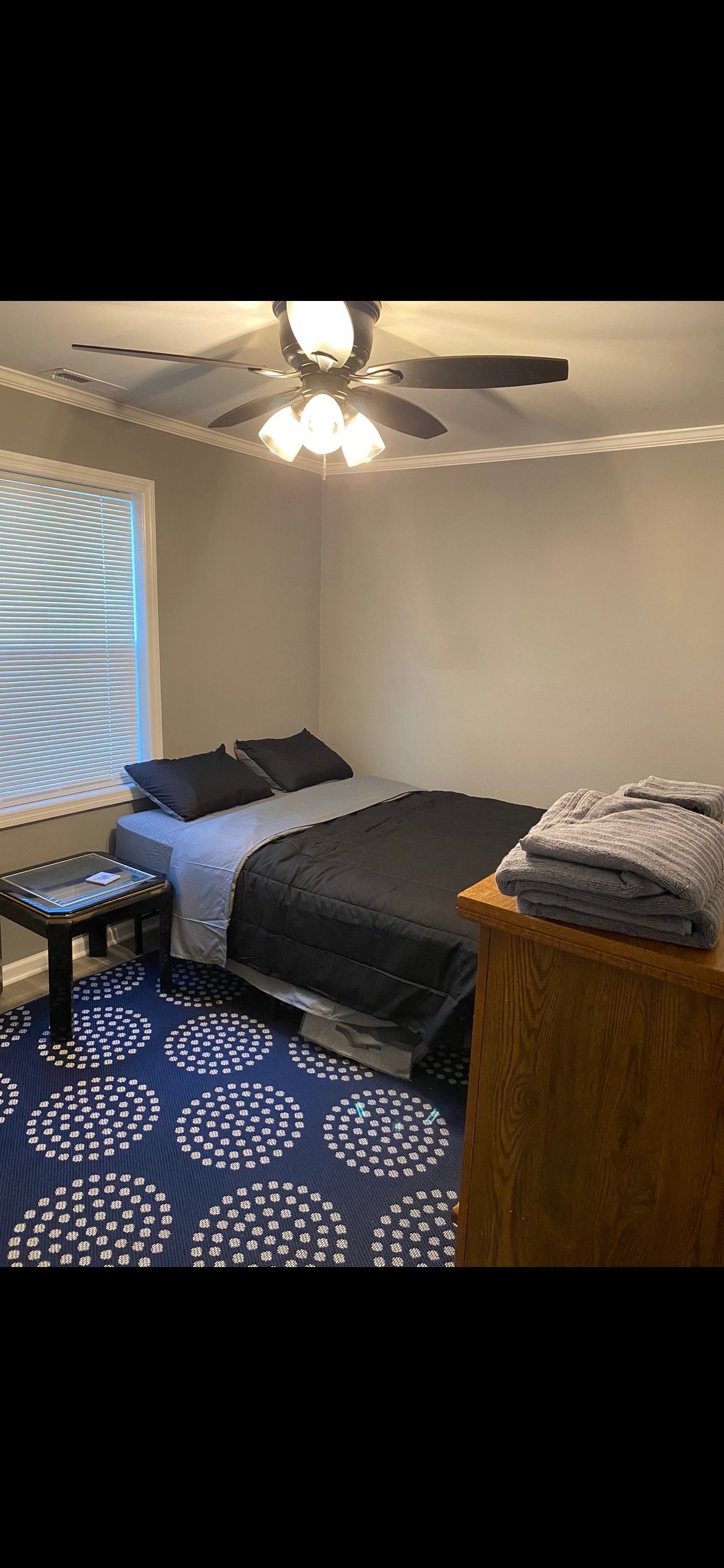 A comfortable bedroom features a spacious bed covered with a dark blanket and two matching pillows. A side table holds a small lamp, and a stack of neatly folded towels is placed on a wooden dresser. Natural light filters through the window, enhancing the room's calm ambiance.