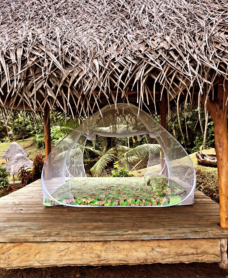 A sheltered outdoor area features a transparent tent on a wooden platform. The tent showcases a vibrant floral-patterned floor mat. Surrounding greenery and tropical plants create a serene environment, while a thatched roof provides protection from the elements.