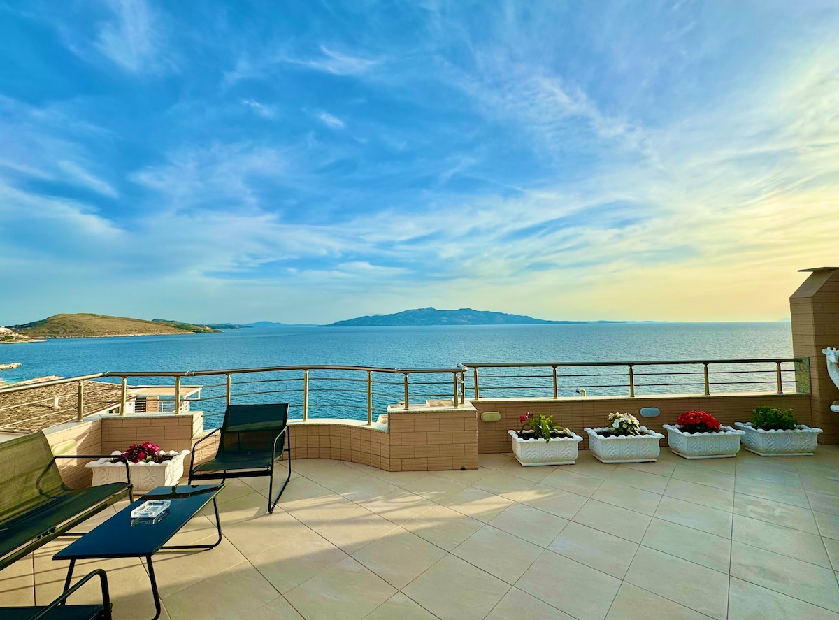 A spacious terrace overlooks a tranquil sea view, featuring comfortable seating options and decorative flowering planters. The expansive patio offers a serene outdoor area, with gentle waves in the distance and a clear sky enhancing the atmosphere.