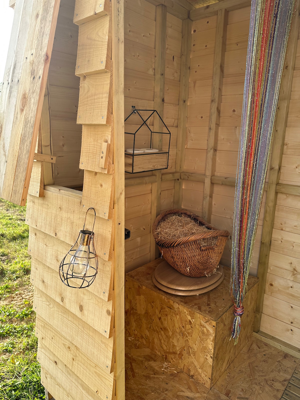 A wooden structure with natural textures and a warm, inviting atmosphere. Inside, there is a woven basket and a round wooden stand on the floor, with delicate items displayed on a small shelf. Colorful strings hang from the side, adding a touch of character.