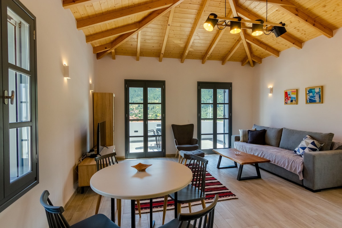 A spacious living area is showcased with a wooden ceiling featuring exposed beams. Light floods the room through multiple windows, highlighting a comfortable sofa and a coffee table. A dining table with chairs is positioned nearby, and decorative art pieces are displayed on the walls.