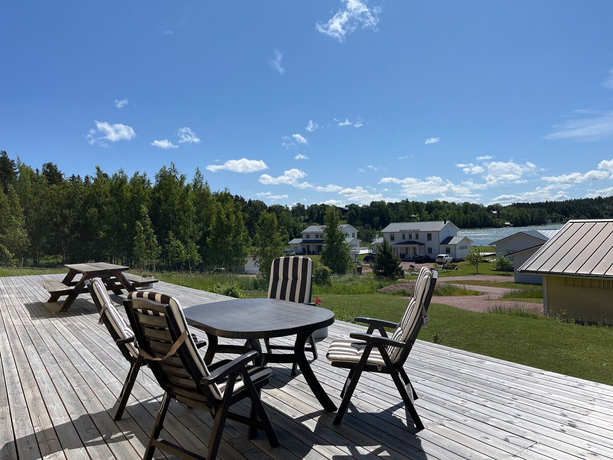 A spacious wooden deck is featured, equipped with a round table and several comfortable chairs. Lush greenery is visible in the background, along with a view of neighboring buildings and the surrounding landscape under a clear blue sky.
