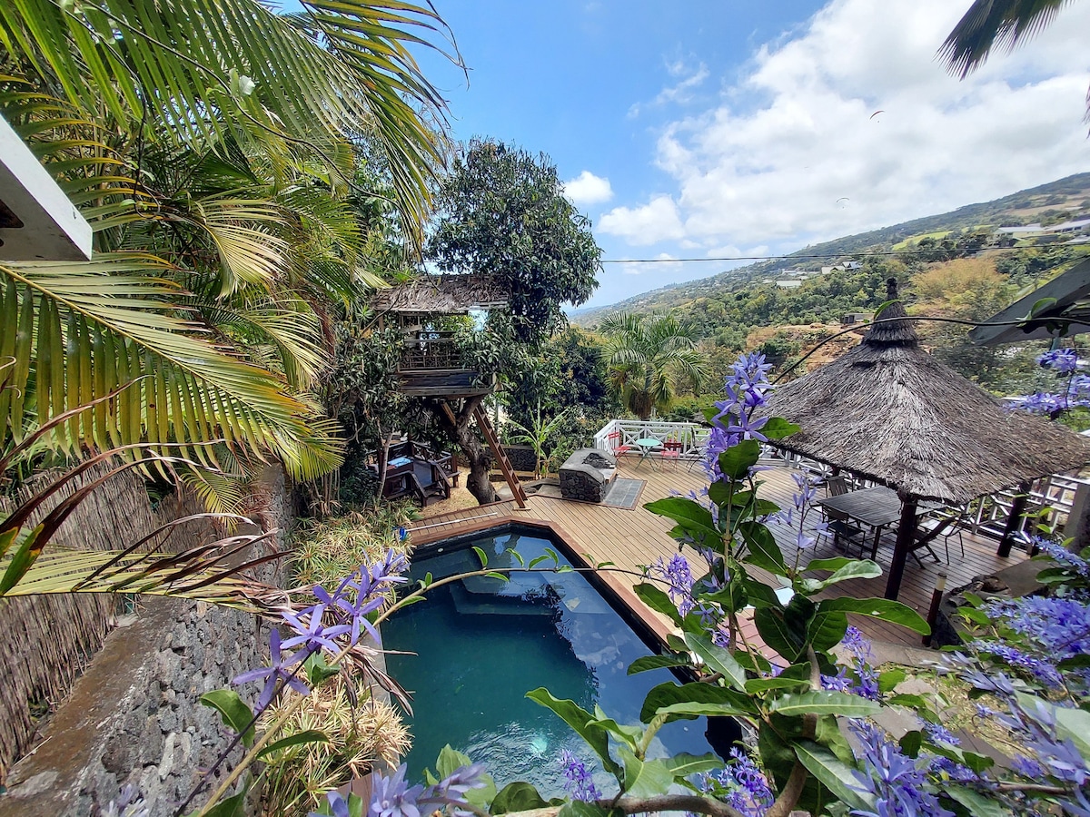 A private pool area is surrounded by lush greenery, with a wooden deck and two thatched-roof gazebos. A treehouse is visible among the trees, providing a playful element to the outdoor space. Vibrant flowers add colorful accents to the scene, enhancing the garden ambiance.
