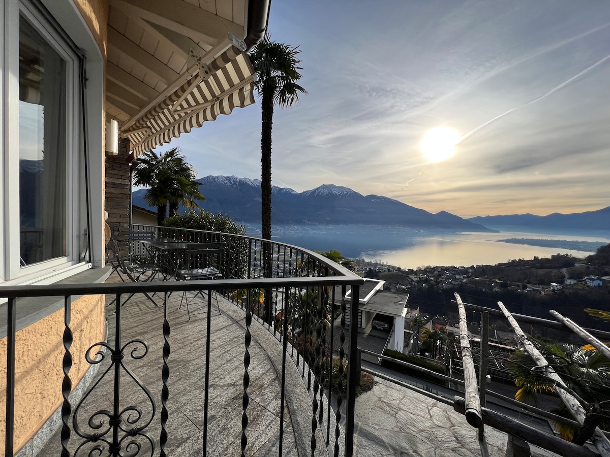A wide balcony is seen with a decorative railing, overlooking Lake Maggiore and mountains in the distance. The sun is positioned low in the sky, casting a soft glow over the landscape. Palm trees are visible on the balcony, adding to the serene setting.