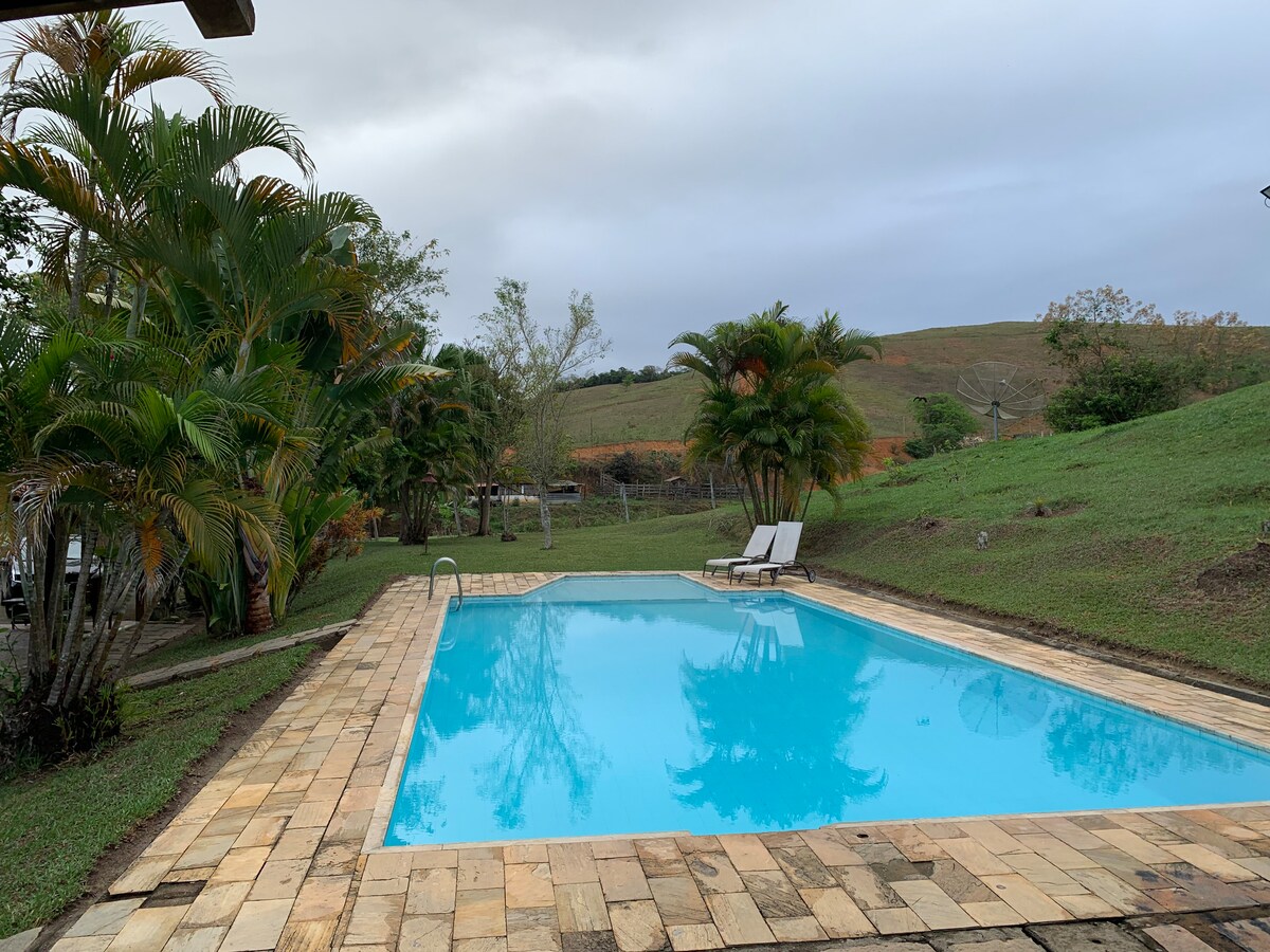 A swimming pool with clear blue water is surrounded by a stone patio, featuring lounge chairs for relaxation. Lush greenery, including palm trees and grass, adds a natural touch, while a gentle slope in the background offers a view of the serene landscape.