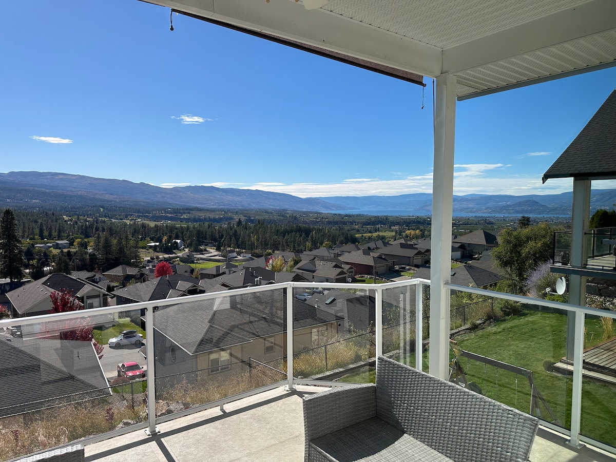A spacious balcony offers a view of the surrounding Black Mountain neighborhood, featuring rolling hills, distant mountains, and a clear blue sky. Comfortable outdoor seating is positioned nearby for relaxation, with the glass railing providing unobstructed visibility of the landscape.