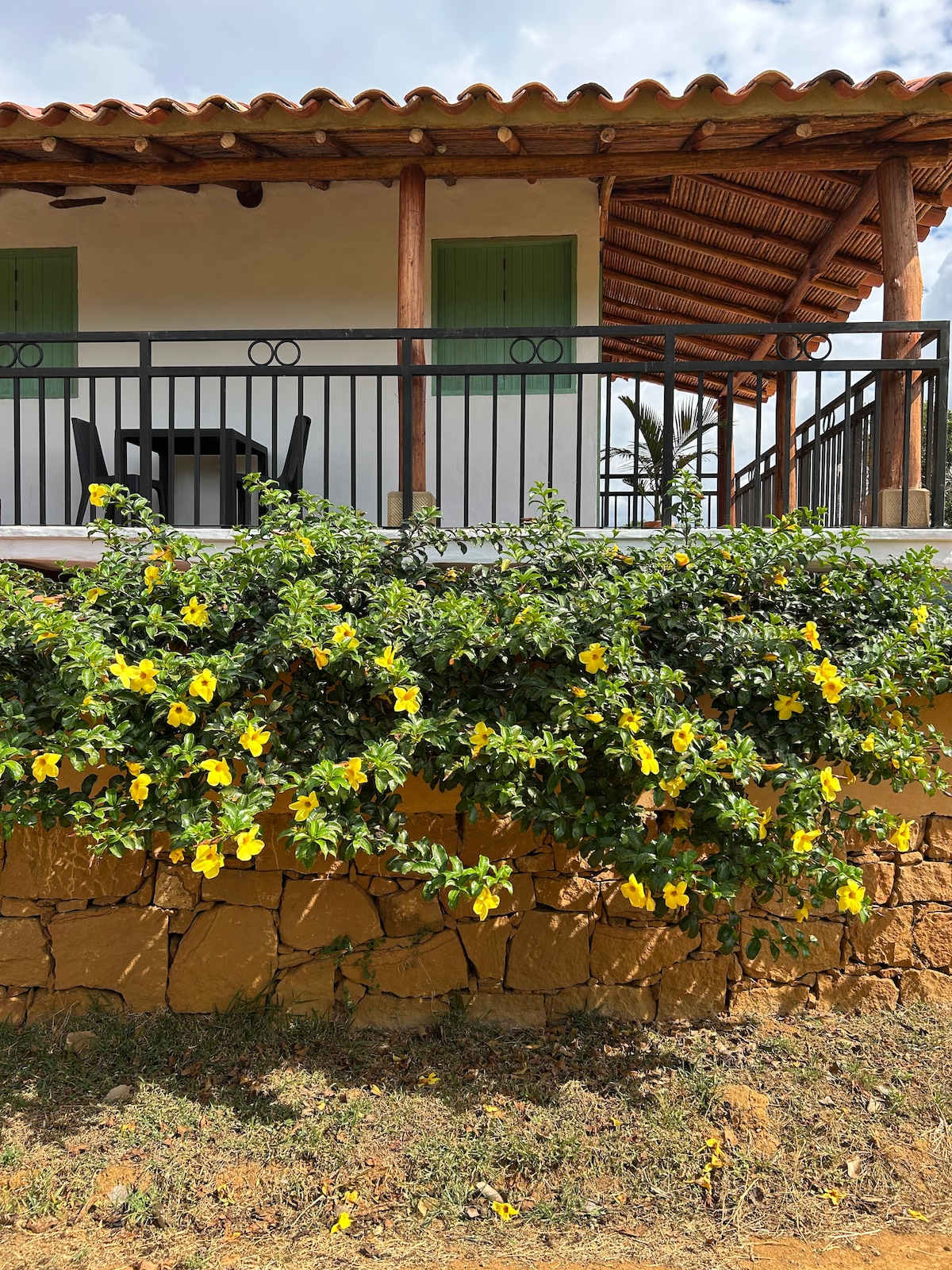 The exterior of a charming cabin features a wooden porch with green shutters and a table and chairs for outdoor seating. Bright yellow flowers cascade over a stone wall, complementing the natural surroundings.
