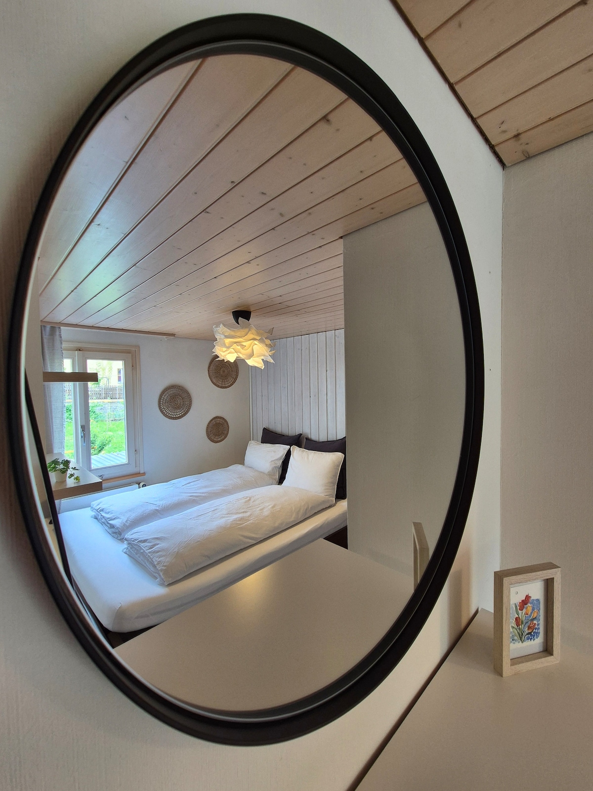 A round mirror reflects a cozy bedroom featuring a spacious bed with crisp white linens. Natural light filters through a nearby window, highlighting textured wall accents. A minimalist bedside table displays a small framed artwork, contributing to a serene atmosphere.