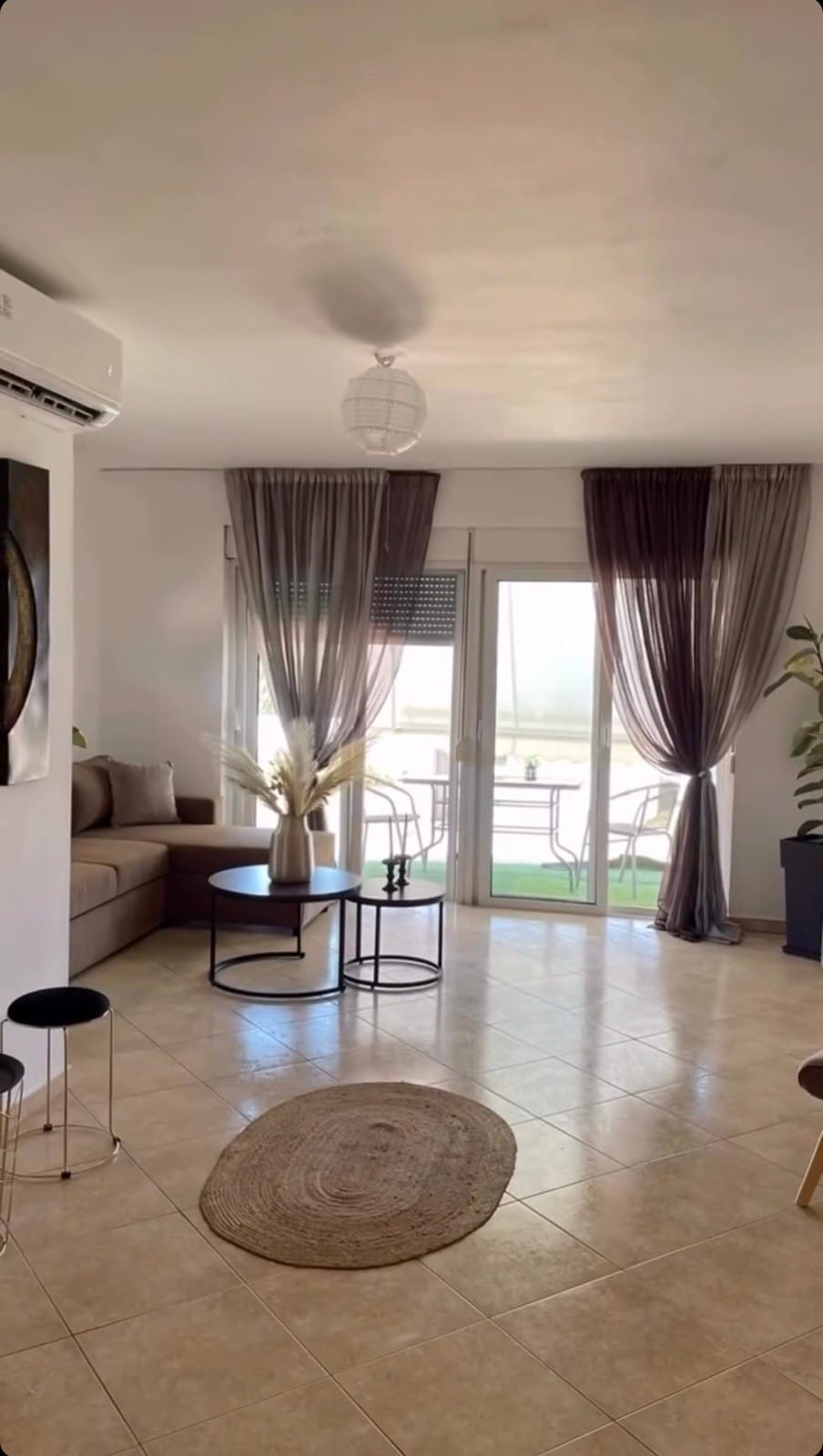 A bright living area is highlighted by large glass doors that open to a balcony. The space features a comfortable sofa, a round coffee table, and decorative plants. Sheer curtains softly filter natural light, while a woven rug adds texture to the tiled floor.