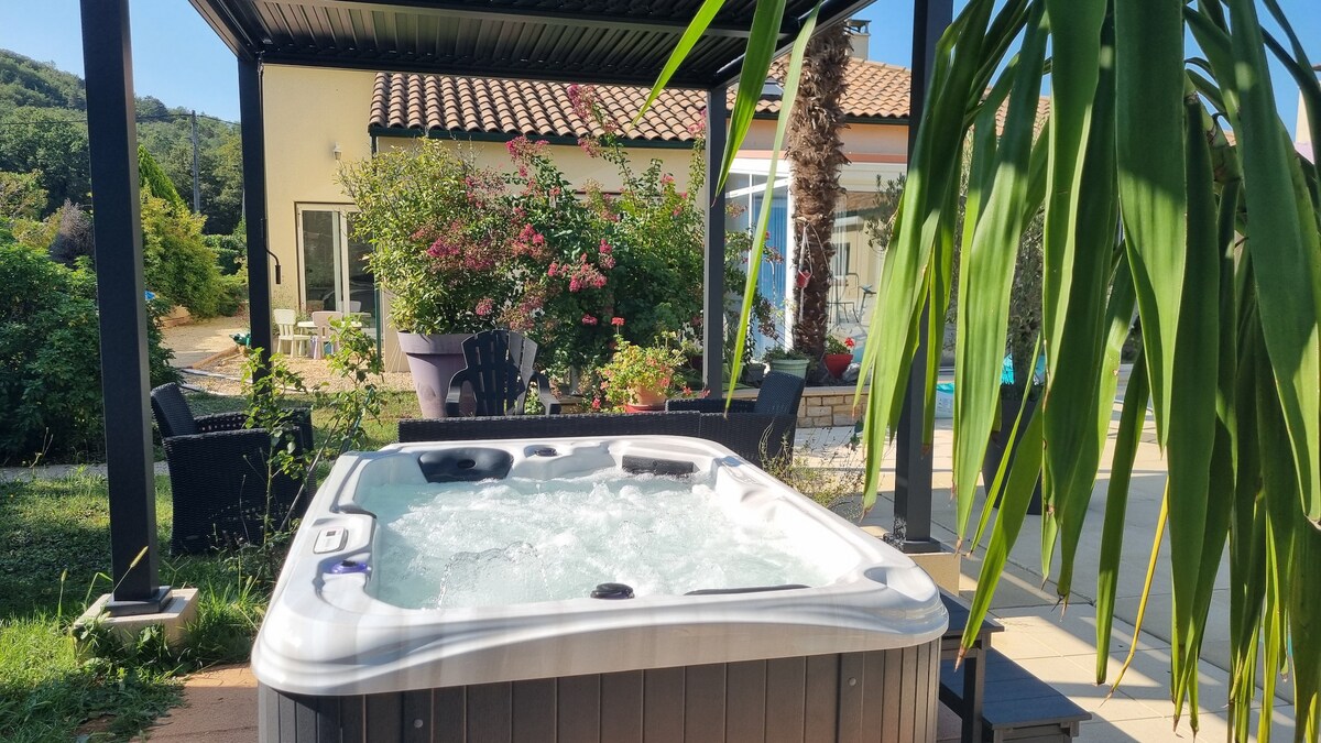 A hot tub is positioned beneath a shaded pergola, surrounded by lush greenery. Colorful flowers and shrubs enhance the outdoor setting, with a distant view of the house showcasing a tiled roof and large windows. Comfortable outdoor seating options are nearby.