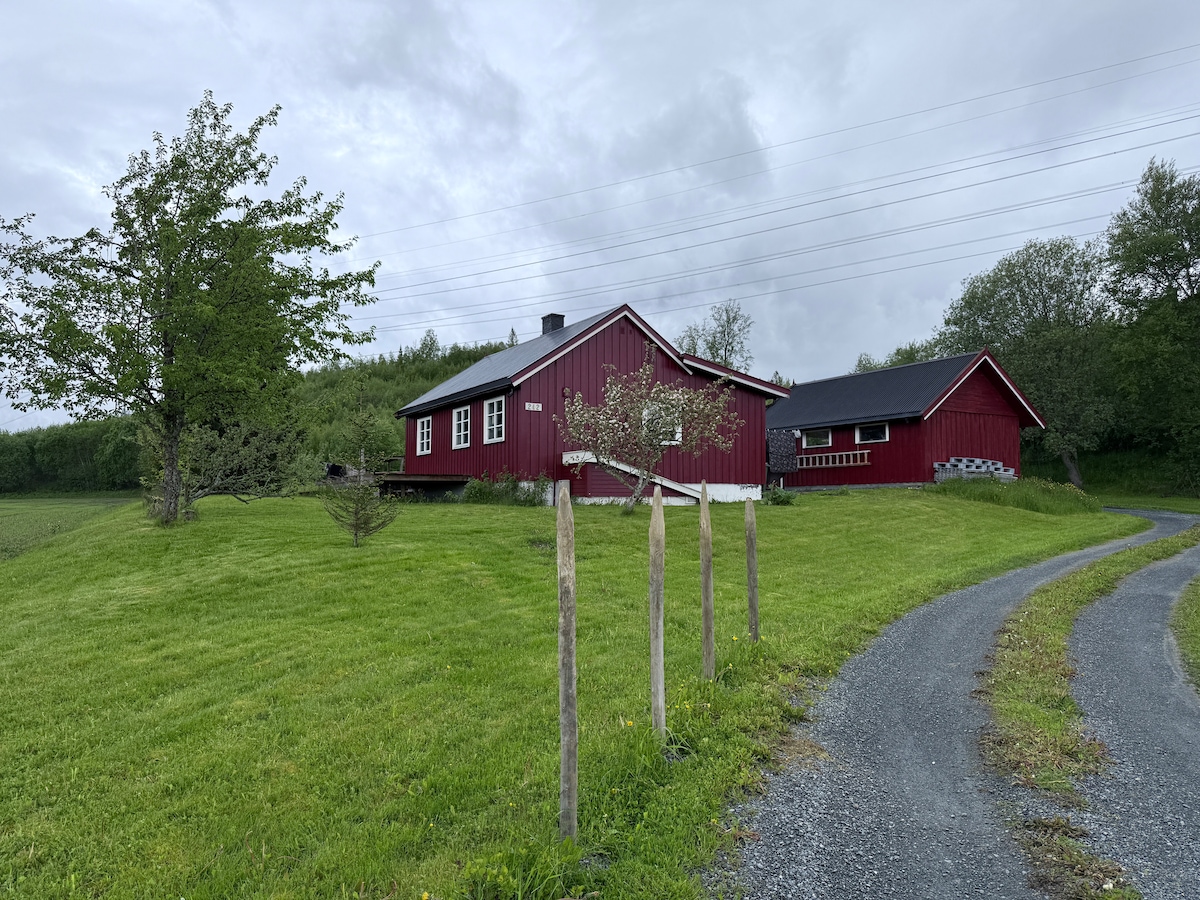 A small, cozy red house is positioned on a gentle hill, surrounded by lush green grass. A winding gravel driveway leads up to the entrance, framed by simple wooden posts and trees in the background. Overcast skies give a serene, calm atmosphere.