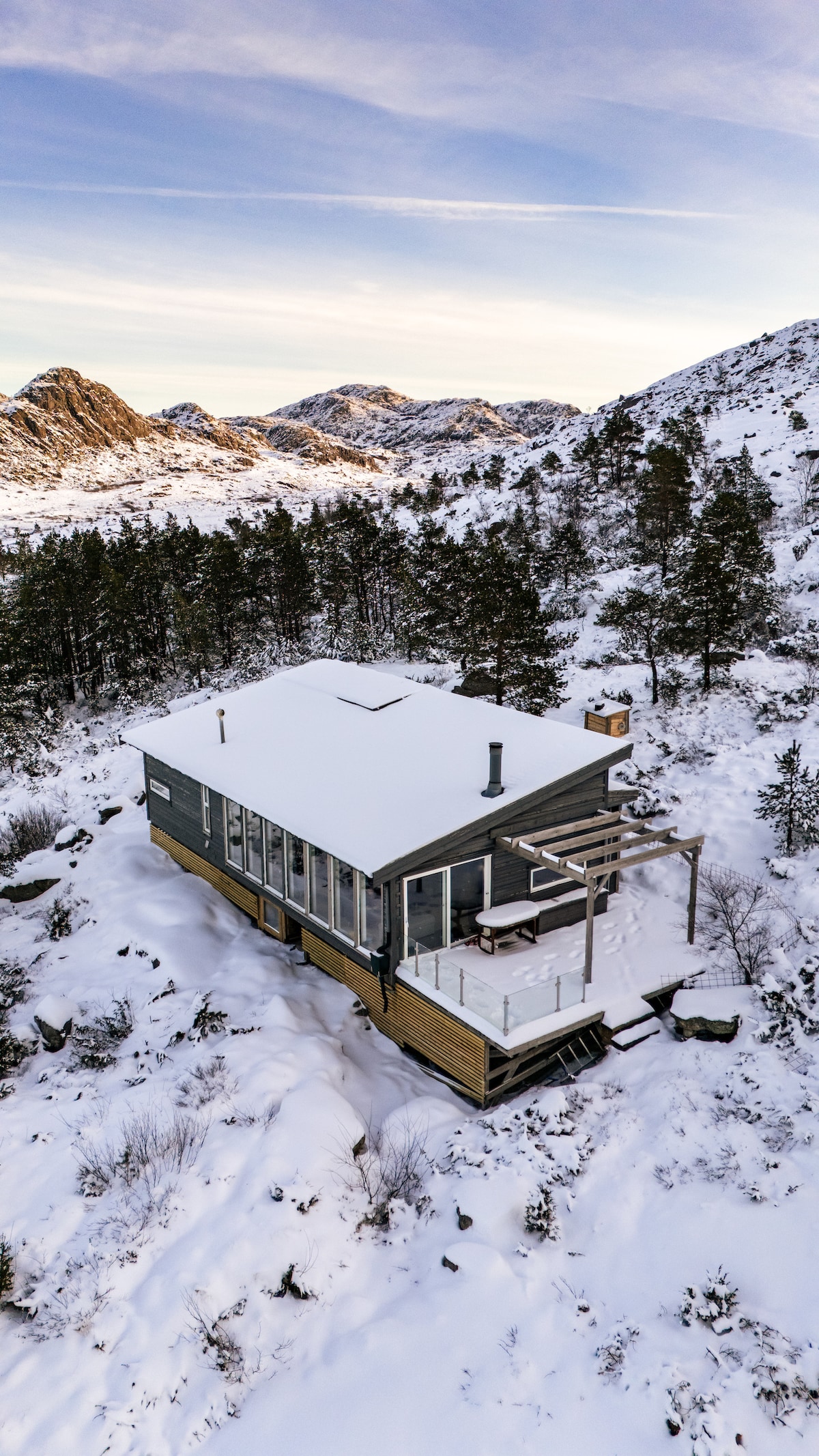 The cabin is surrounded by a serene snowy landscape, with a sloped roof blanketed in white. It features large glass windows, providing views of the mountainous terrain, while a small deck extends outward, adding to the connection with nature.