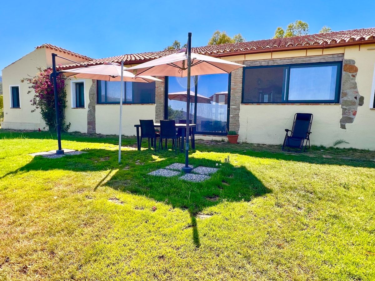 An outdoor seating area is seen beneath two spacious awnings, providing shade for a table with six chairs. The green lawn and natural surroundings enhance the inviting atmosphere of this relaxing space. Large windows of the villa reflect the sunlight, contributing to a cheerful ambiance.