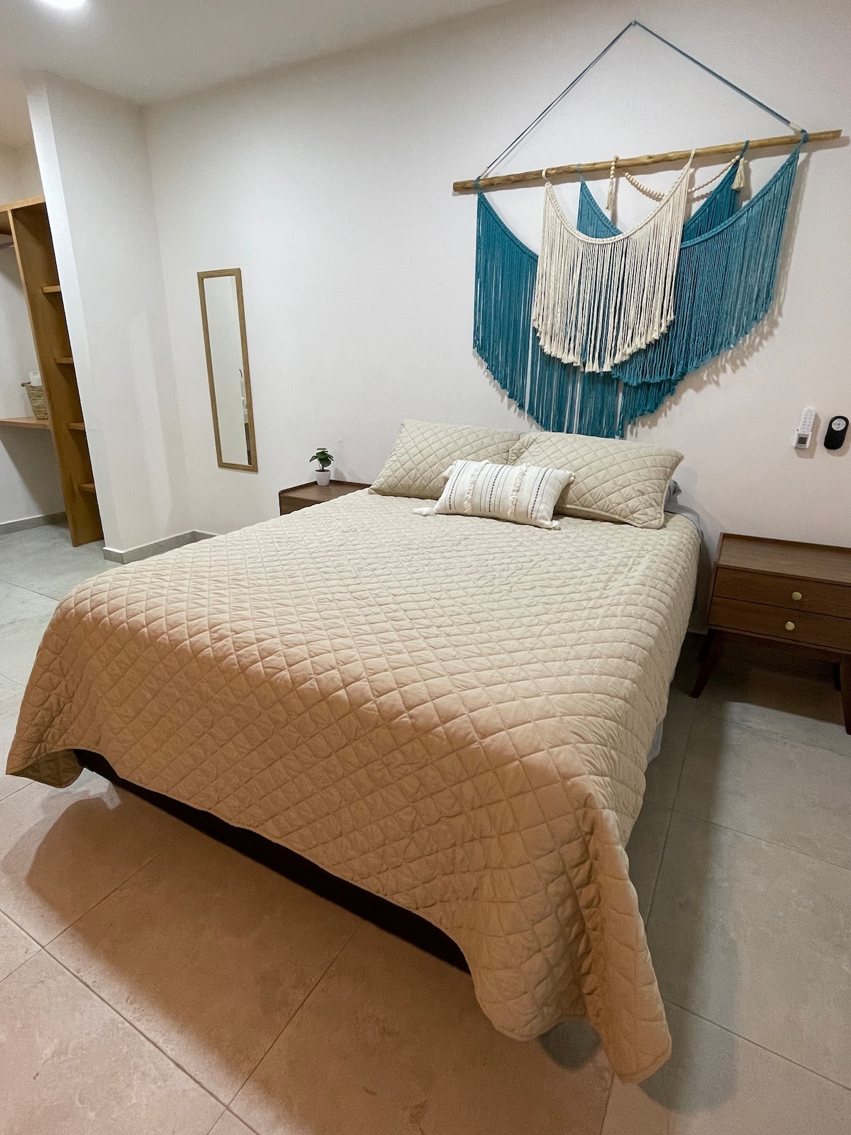 A spacious sleeping area features a queen-size bed adorned with a light quilt. On either side, wooden nightstands hold decor. A woven wall hanging adds texture, while a mirror reflects the simplicity of the room. Neutral tones create a calming environment.