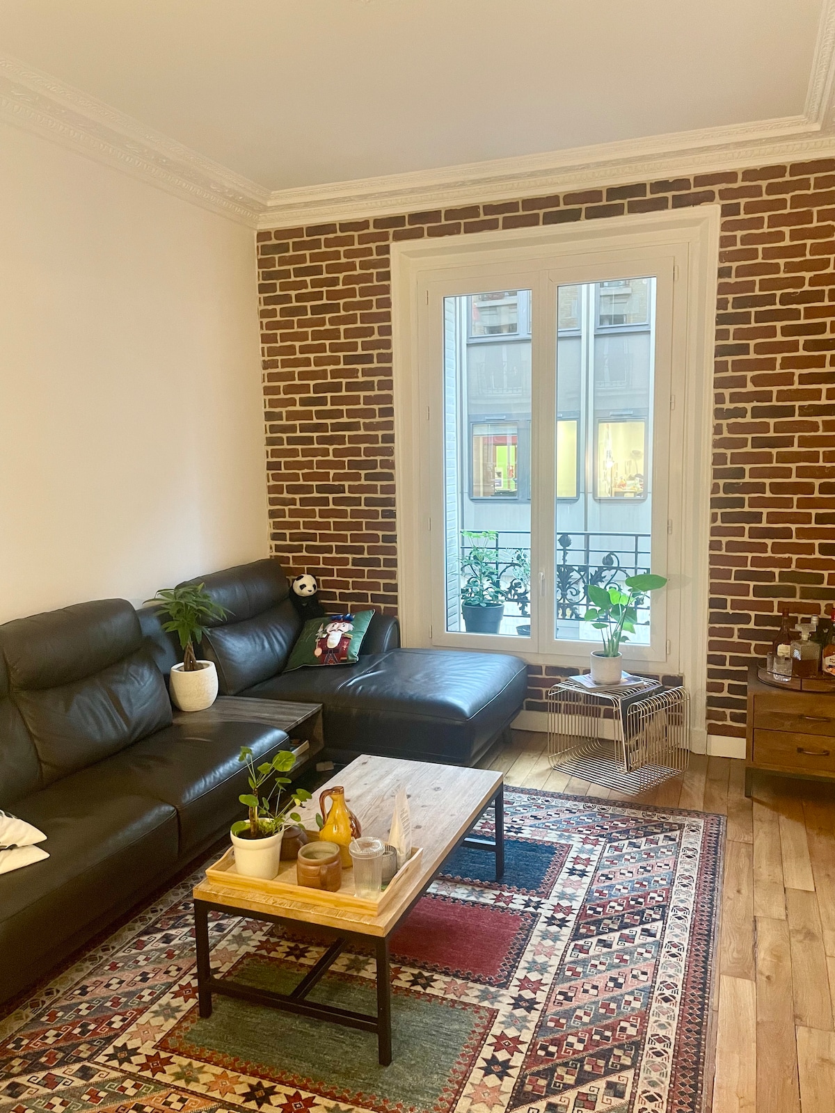 A living area features a dark leather sectional sofa complemented by an ottoman. A coffee table adorned with small plants and decorative items sits centrally on a patterned rug. A large window provides natural light and overlooks an external view.
