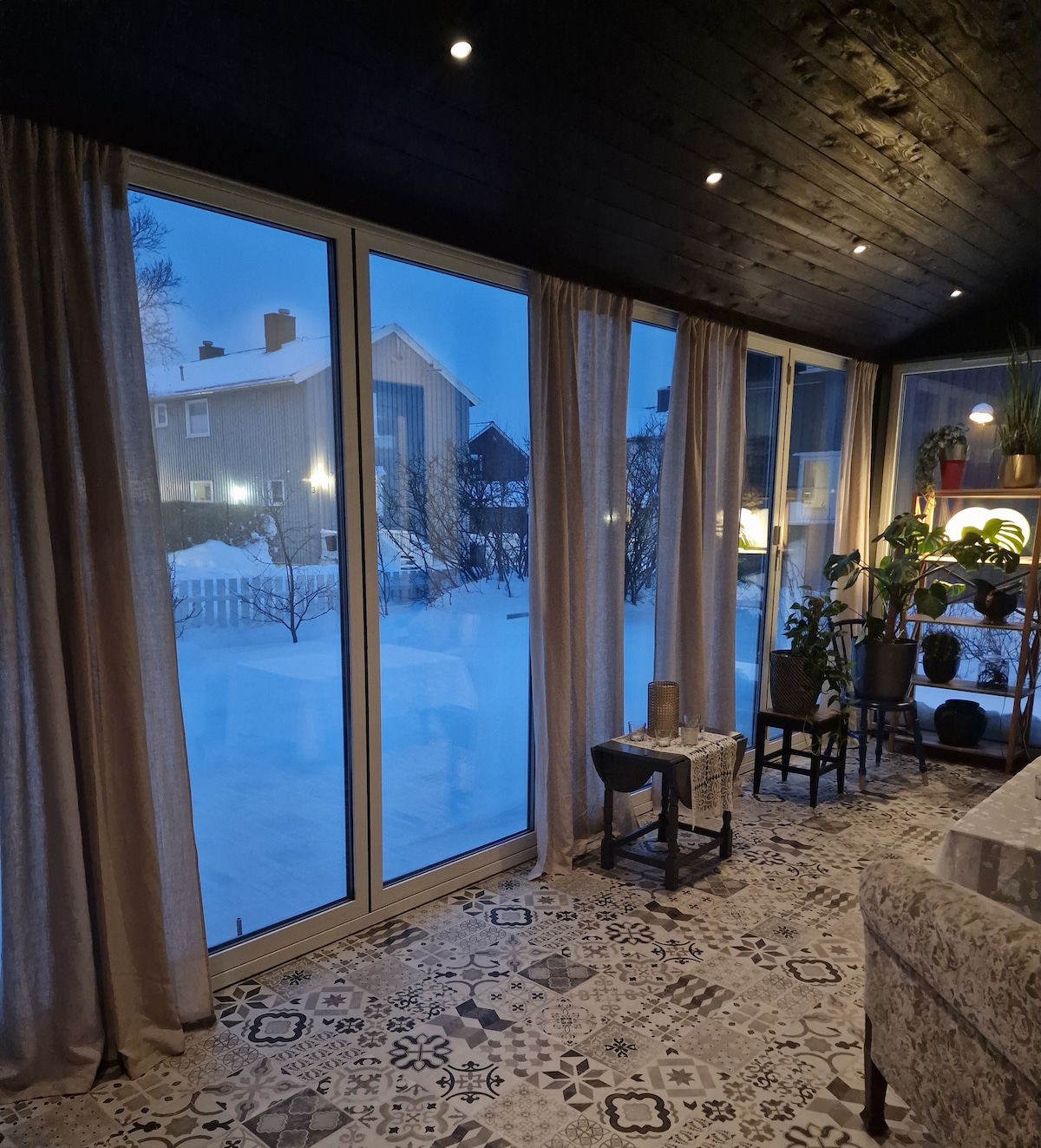 A spacious sunroom features large glass windows, providing a view of the snowy landscape outside. Soft curtains frame the windows, while patterned flooring enhances the interior. Potted plants are placed strategically, adding a touch of greenery to the space.