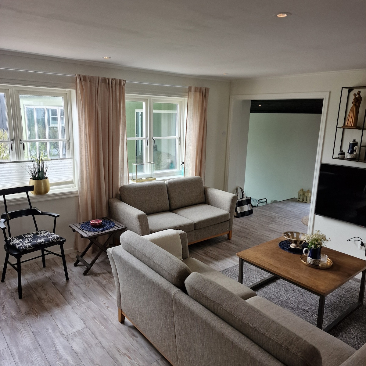 A comfortable living area is showcased, featuring two soft sofas and a wooden coffee table. Natural light enters through multiple windows adorned with light curtains. A small plant sits on the table, complementing a minimalistic decor. A cozy chair and a small side table are also visible.