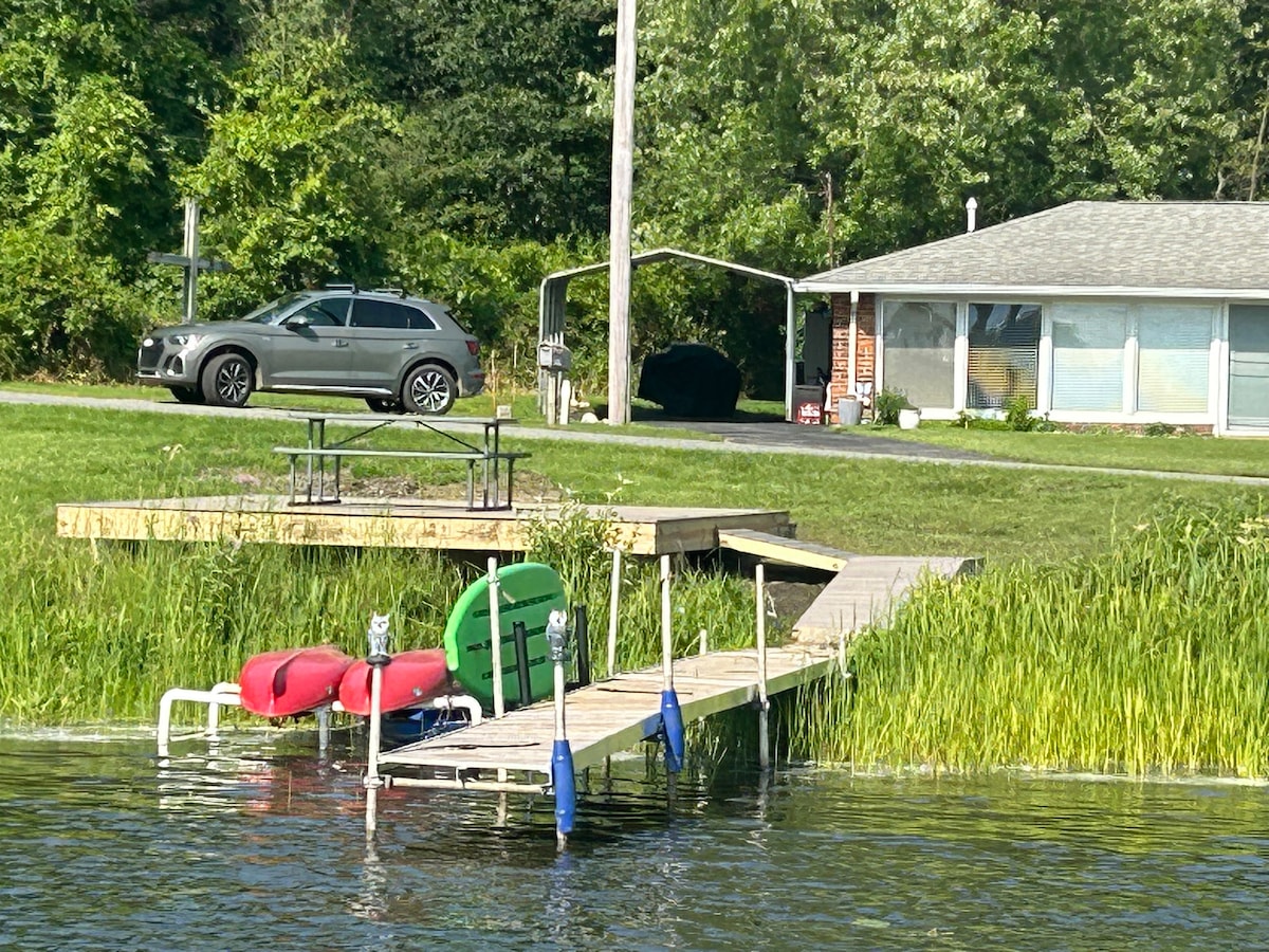 A tranquil lakeside view features a wooden dock extending into the water, equipped with two kayaks and a paddleboard. In the background, a single-story home is visible with an adjacent carport and picnic table on a grassy area.