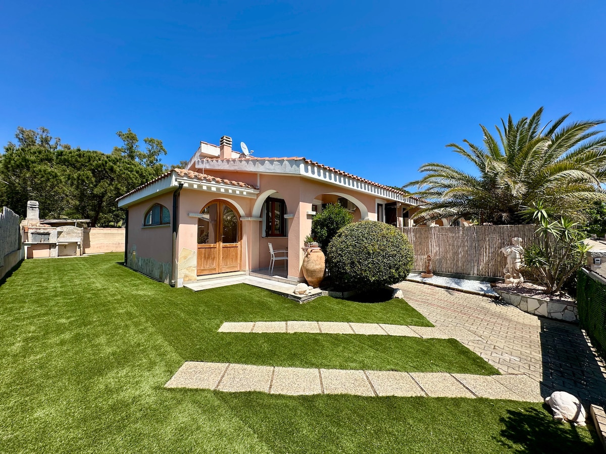 A villa is surrounded by a lush garden with manicured grass and decorative pathways. The exterior features a charming facade with arched windows and a wooden door. A palm tree and other greenery add to the tranquil atmosphere. A stone path leads to the entrance.