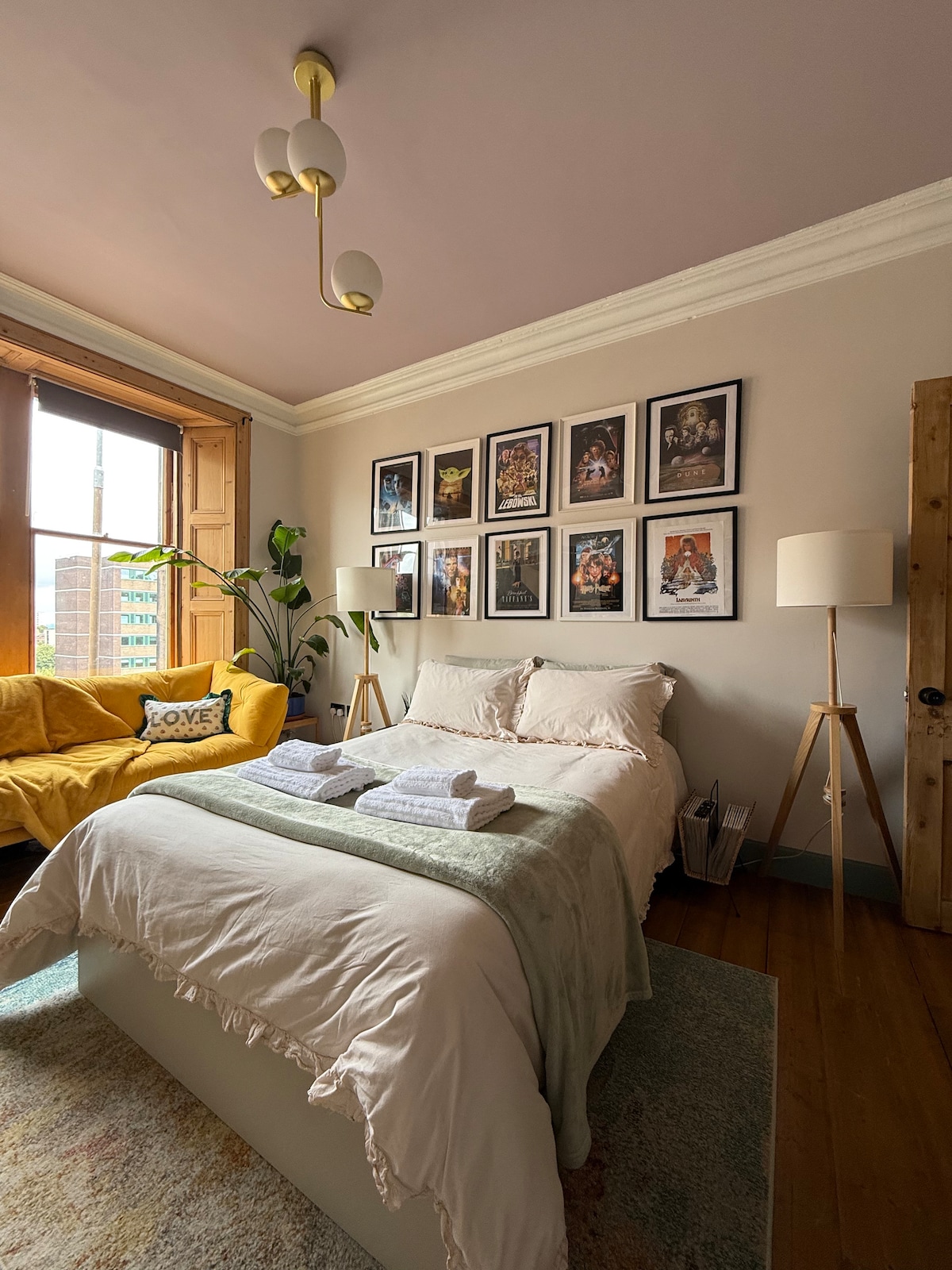 A cozy room features a spacious double bed dressed with soft linens and a light blanket. Framed artwork is displayed on the wall above, while a yellow sofa is positioned nearby. Natural light filters through a large window, enhancing the warm tones of the interior.