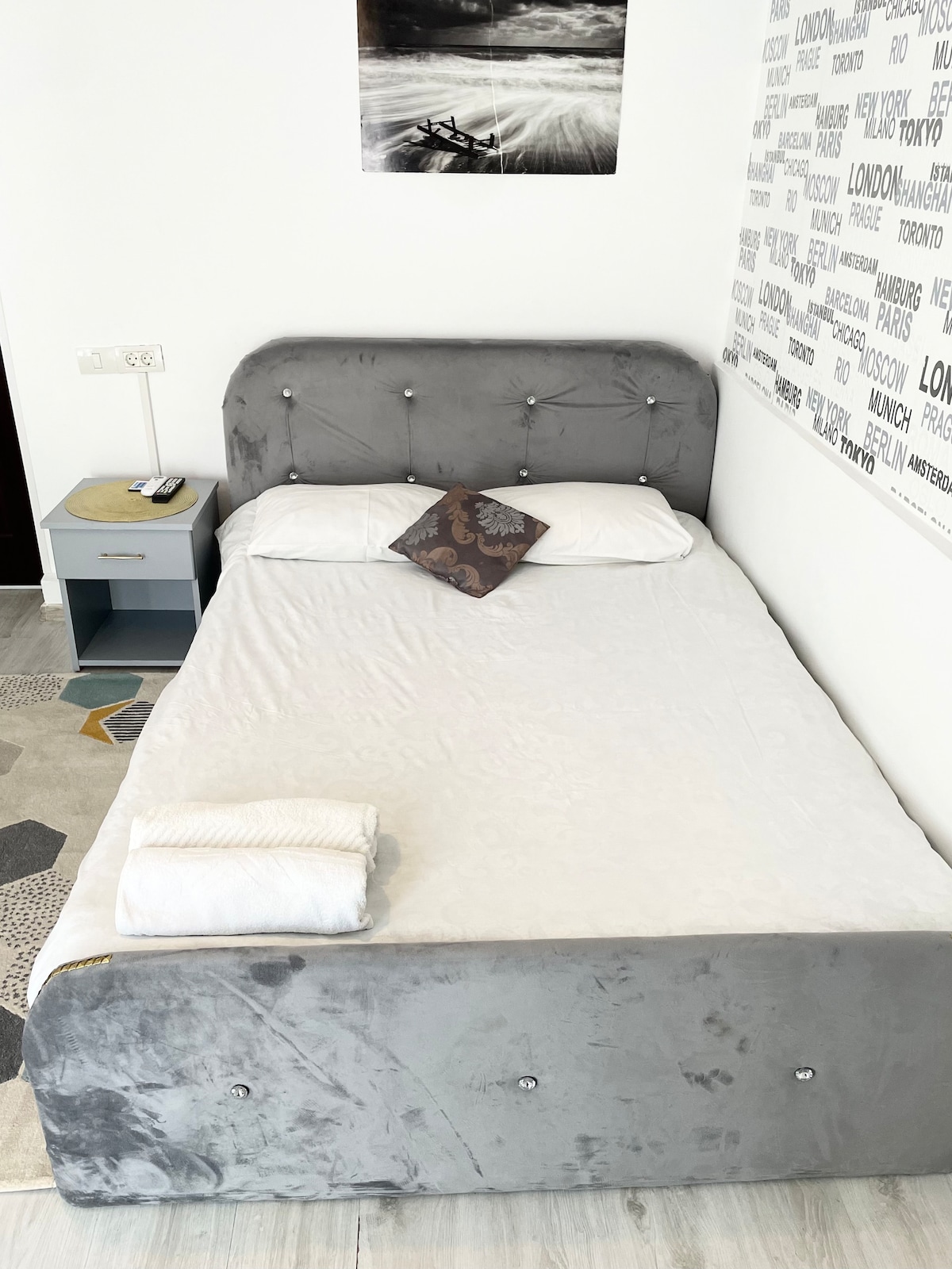A spacious bed dressed in crisp white linens is presented in a well-lit room. A decorative pillow adds a touch of style. Nearby, a small bedside table holds a lamp and a telephone. Light-colored flooring contributes to a clean and modern aesthetic.