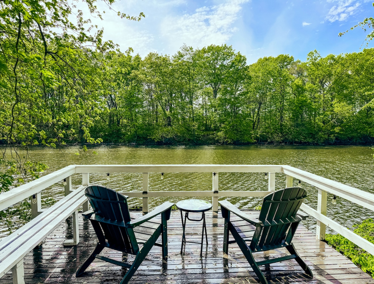 A serene dock overlooks a calm creek surrounded by lush greenery. Two black Adirondack chairs are positioned beside a small round table, creating a peaceful space for relaxation. The sunlight reflects off the water, enhancing the tranquil setting.