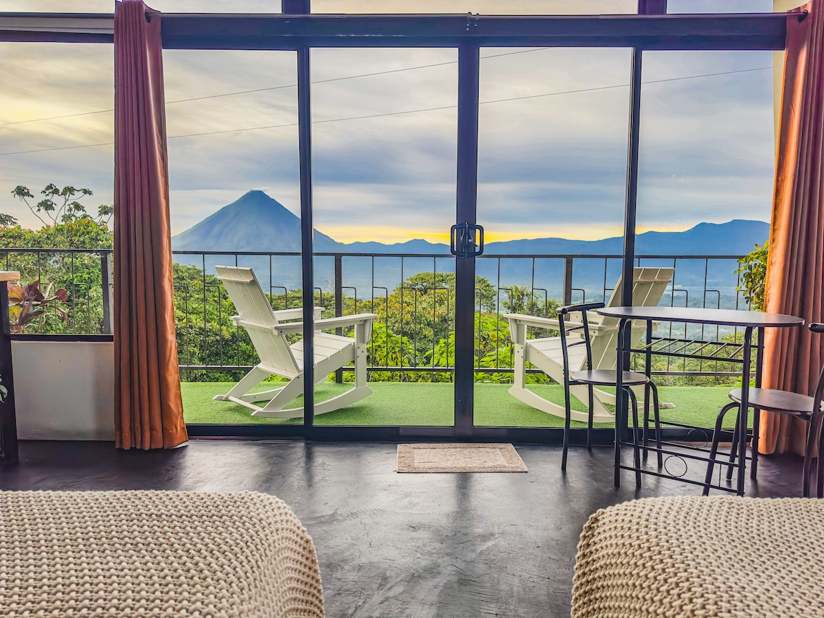 A spacious interior is showcased through large glass sliding doors, revealing a breathtaking view of the Arenal Volcano and surrounding mountains. Two white lounge chairs are positioned on a private balcony, complemented by a small round dining table, inviting moments of relaxation and connection with nature.