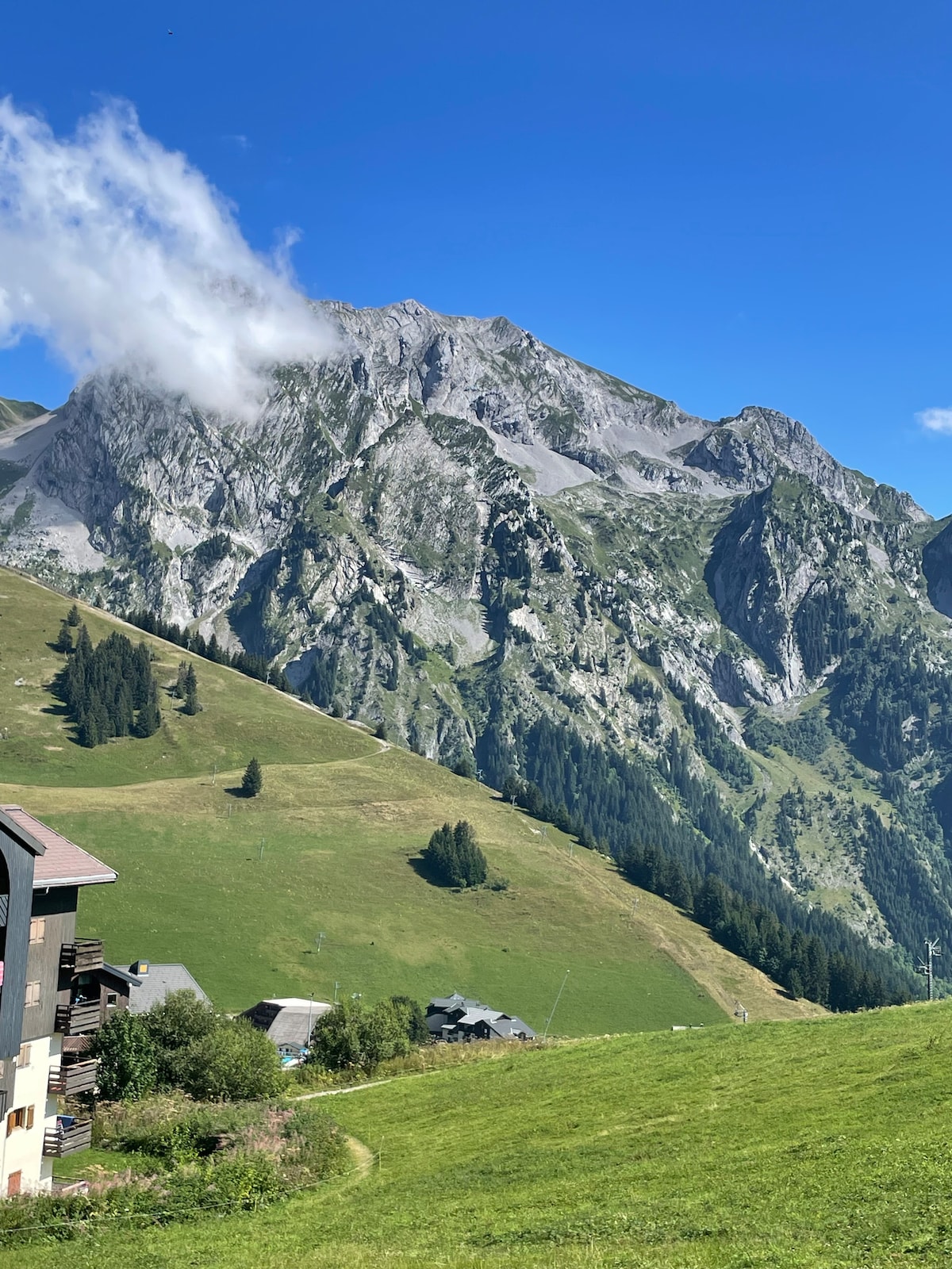A panoramic view of the majestic mountains is displayed, with steep, rocky peaks partially shrouded in wispy clouds. Lush green hillsides lead down to a quaint village nestled among the trees, under a clear blue sky.