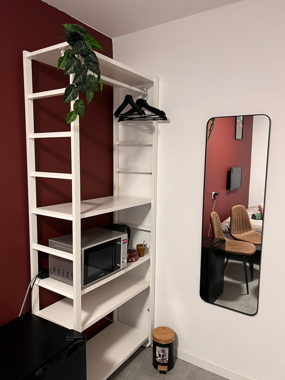 A shelving unit against a deep red wall is featured, showcasing several shelves with hangers and a microwave. A large mirror reflects the space, while a black mini-fridge sits below. The background reveals soft chairs and a television.