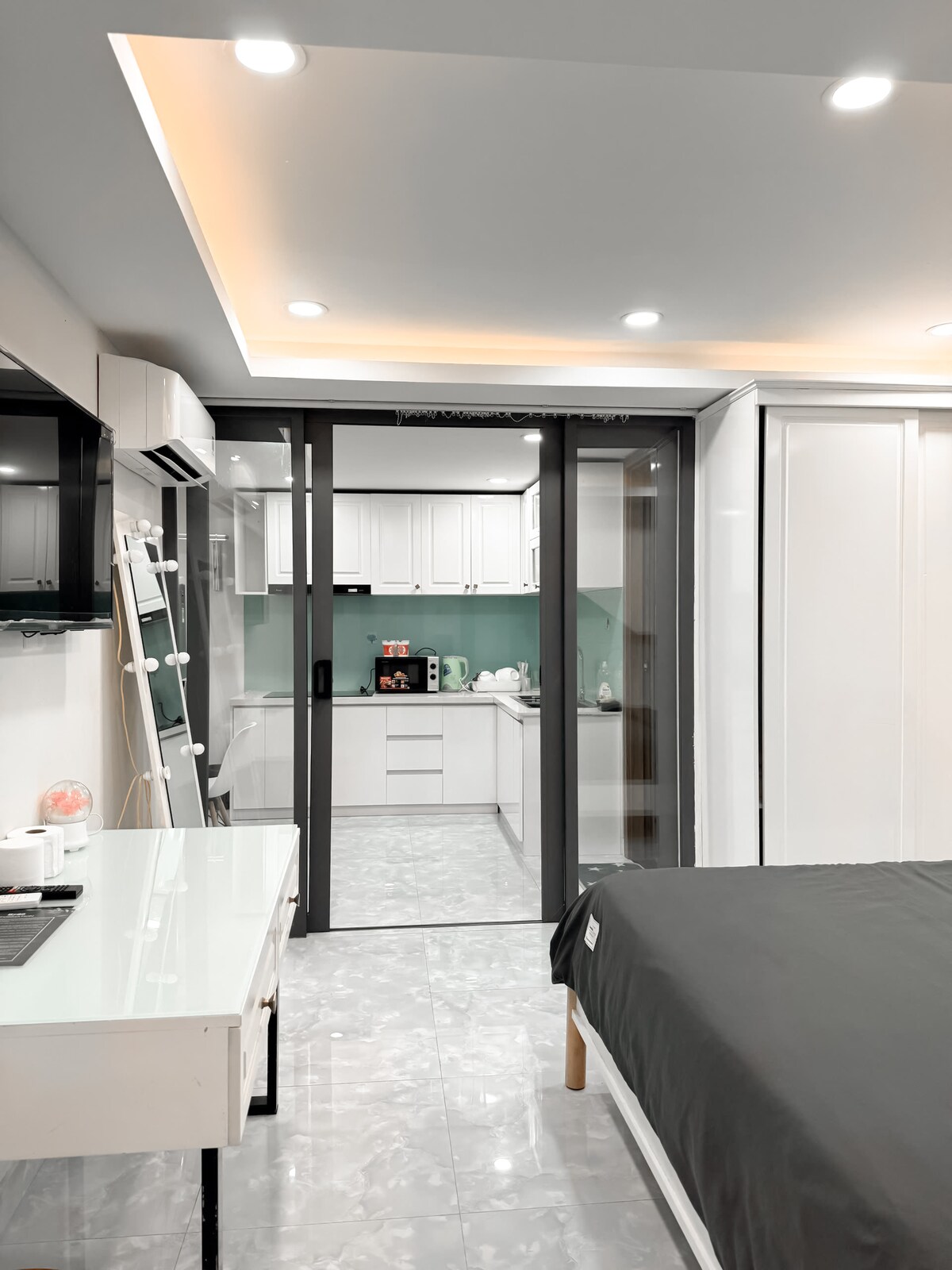 A modern studio room is displayed, featuring a spacious bed with dark bedding. A glass partition reveals a well-equipped kitchenette with a white countertop. Bright overhead lights illuminate the room, while a small desk is positioned nearby, complementing the contemporary decor.