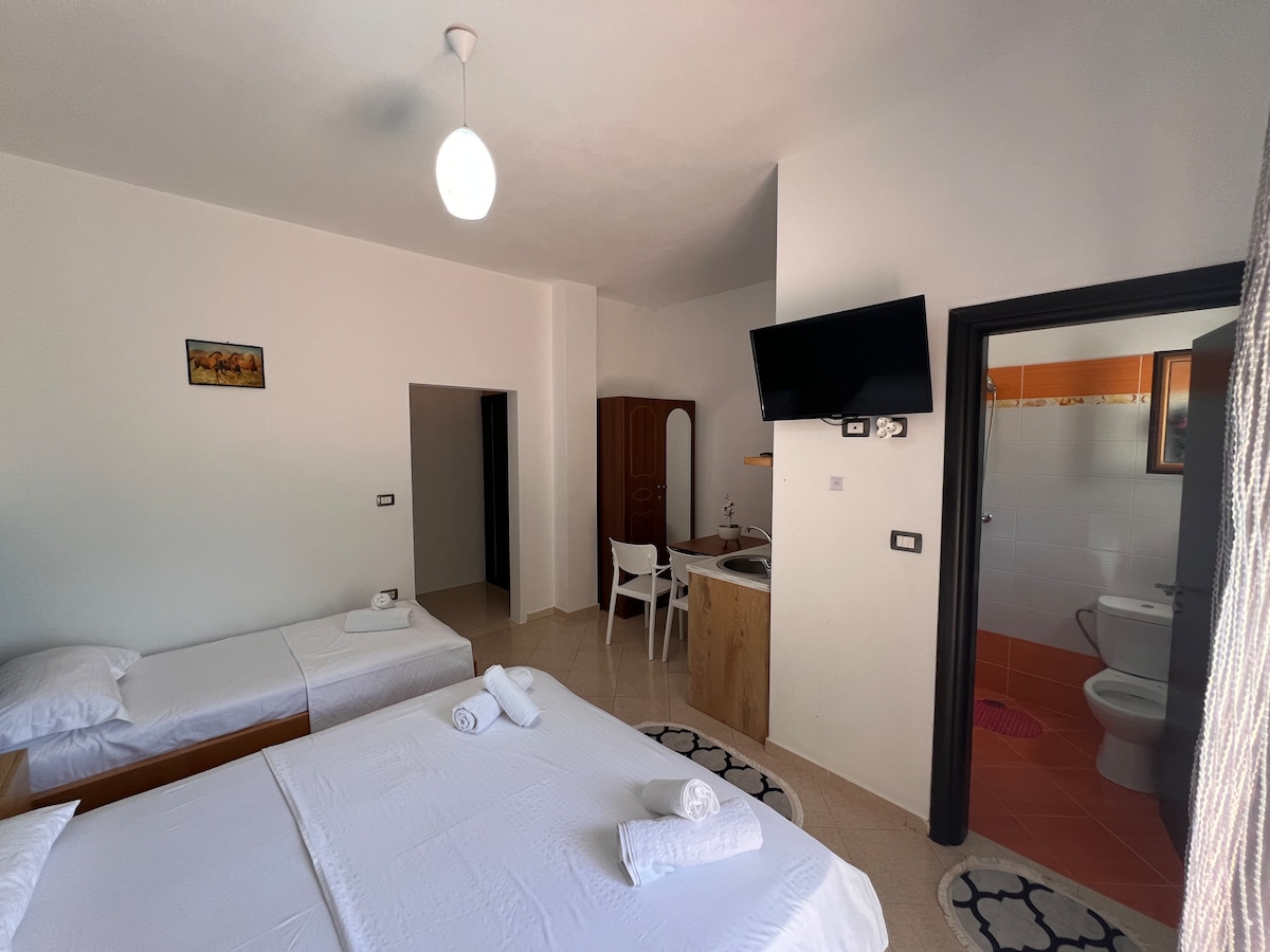 A spacious room is illuminated by natural light, featuring a large bed and a single bed with neatly arranged towels. A wooden wardrobe and a well-placed television enhance the space, while a bathroom area is partially visible in the background.