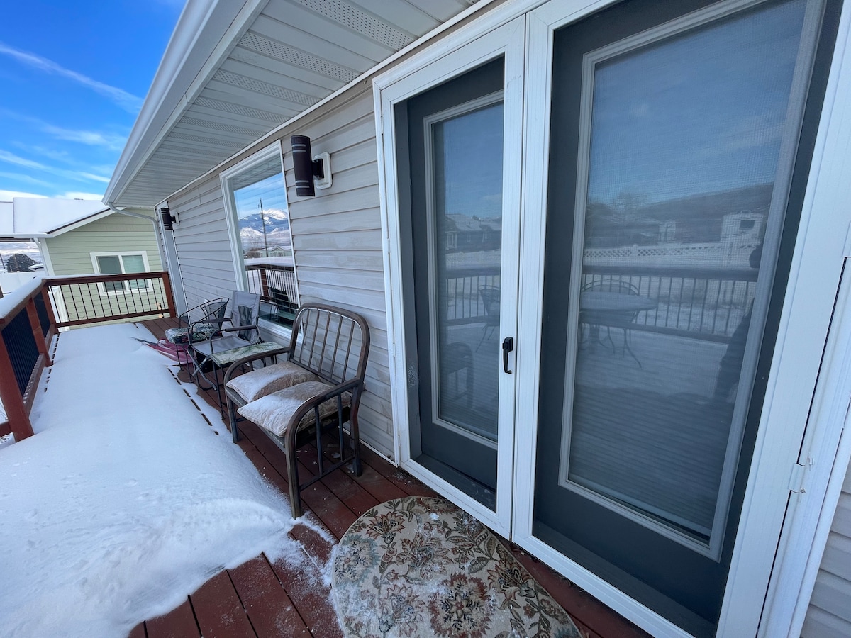 A back patio area is displayed, featuring patio doors leading into the house. Two metal chairs and a small table are visible, surrounded by snow. A patterned rug is on the wooden deck, creating a comfortable outdoor space with views of the surroundings.