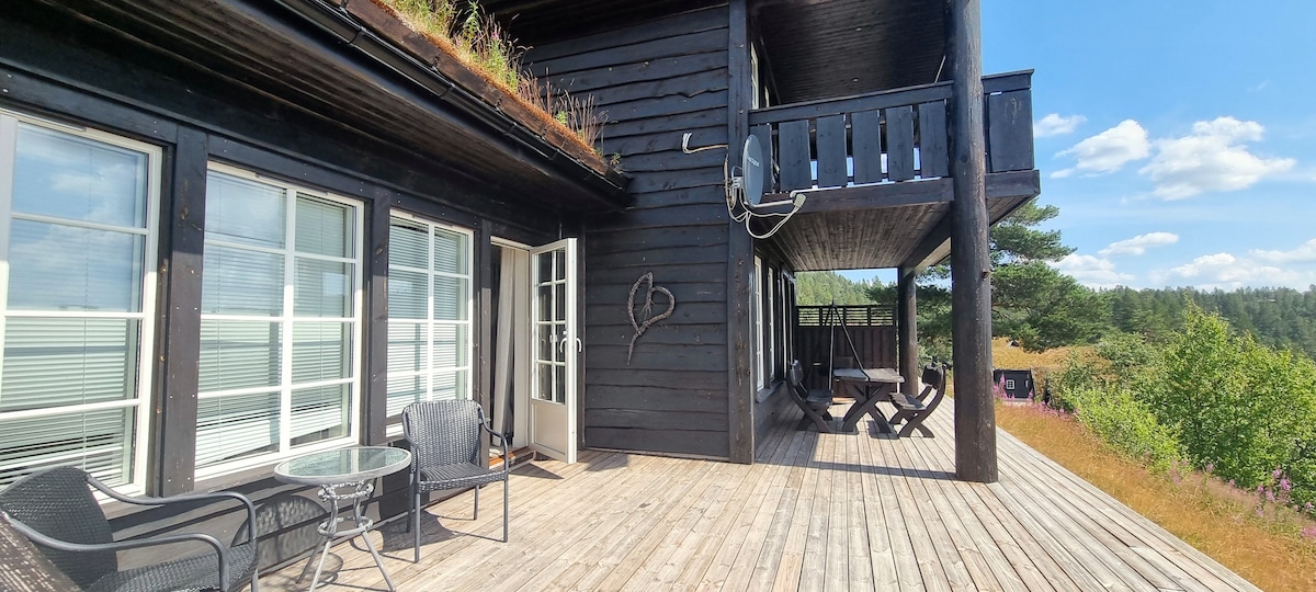 A spacious wooden terrace features seating arrangements including a glass-top table and chairs. An upper balcony can be seen, enhancing the view of the surrounding greenery. Large, multi-paned windows allow natural light to fill the exterior, showcasing the inviting outdoor space.