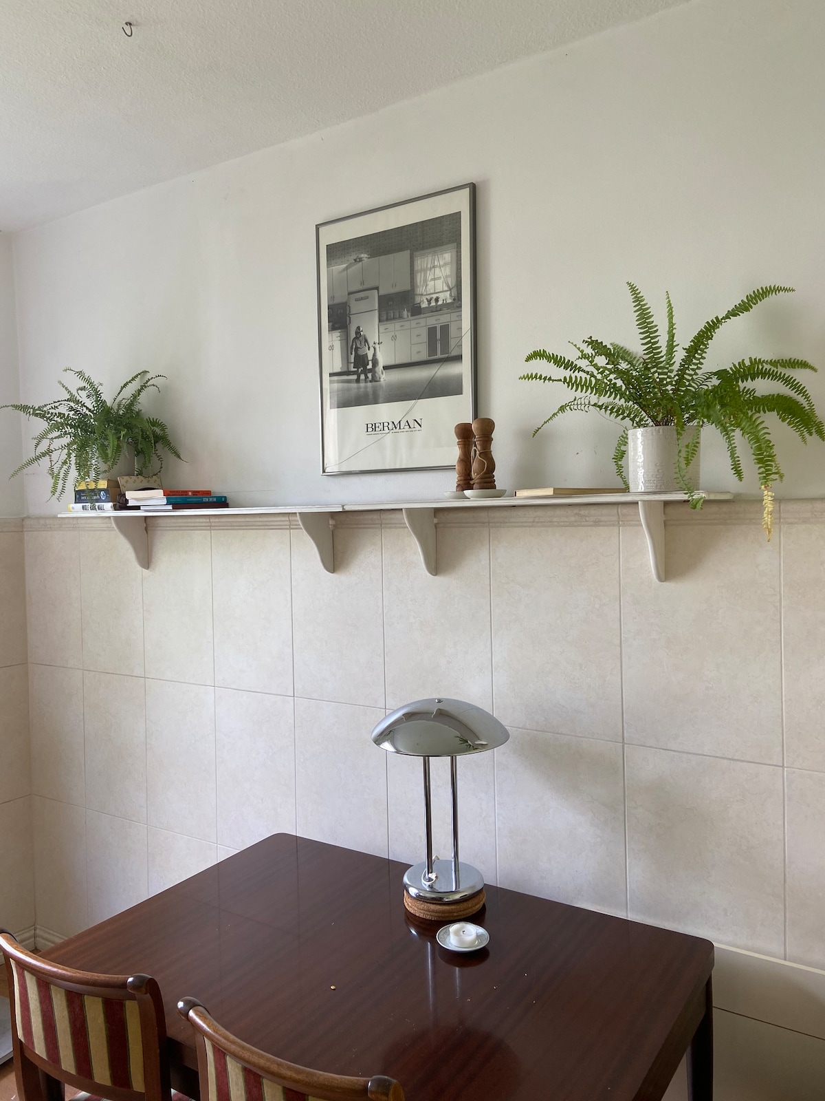 A compact dining area features a wooden table accompanied by two chairs. Decorative shelves above hold books and potted ferns. A silver lamp sits on the table, with a small dish beside it, enhancing the practicality of the space.