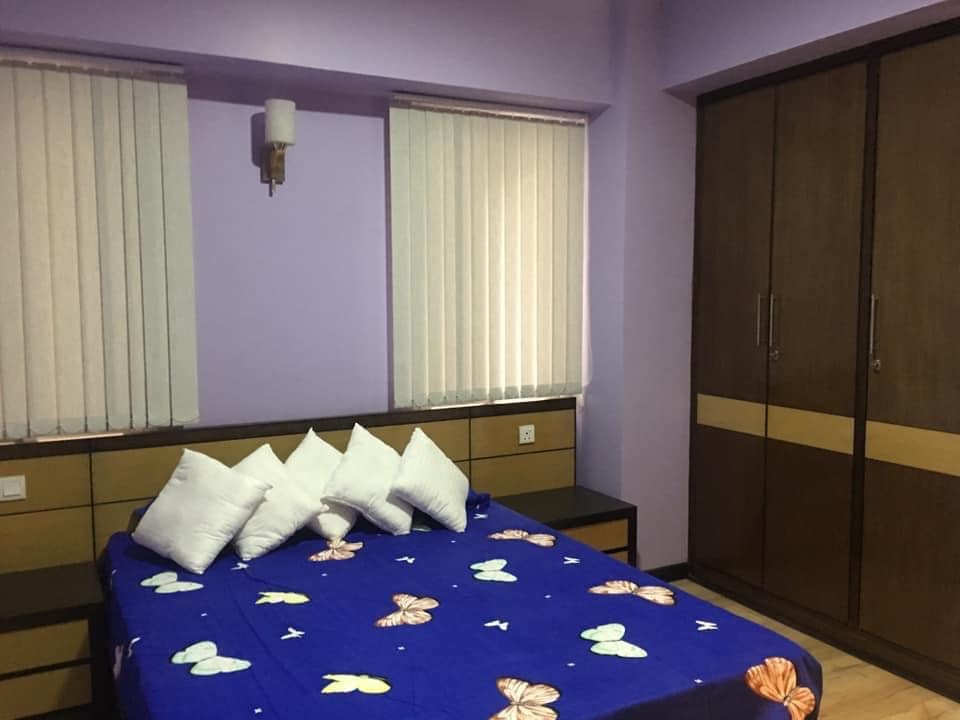 A colored bedspread adorned with butterfly patterns covers a spacious bed, complemented by several white accent pillows. Two windows with vertical blinds allow natural light to illuminate the purple walls, while a wooden wardrobe adds a touch of functionality to the room.
