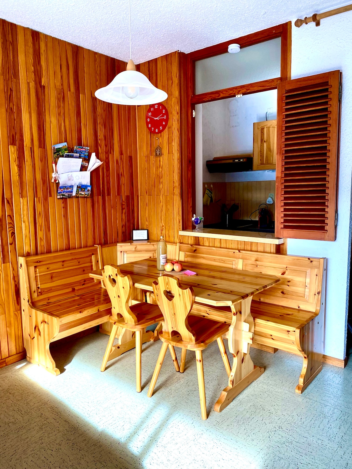 A wooden dining area features a fitted bench and a matching table, providing seating for four. Light streams in through a window with wooden shutters, highlighting the natural grain of the wood and creating a warm, cozy atmosphere.