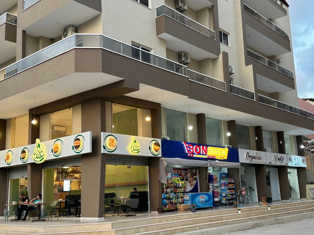 A newly developed building houses various ground floor businesses, featuring a restaurant with outdoor seating and a convenience store. Bright signage indicates the stores, while shaded areas provide a comfortable space for patrons. The modern architecture enhances the vibrant street atmosphere.