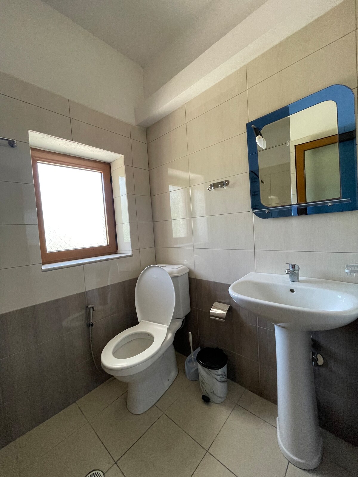 A clean bathroom is displayed, featuring neutral-colored tiles. A window allows natural light to enter, while a mirror above the sink accentuates the space. Essential fixtures include a toilet and a pedestal sink, with a small wastebasket positioned nearby.