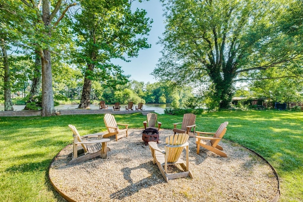 An outdoor fire pit area is surrounded by a circular arrangement of wooden adirondack chairs, set on gravel. Lush green grass and large trees frame the space, with the serene Watauga River visible in the background, and additional seating positioned along the riverbank.