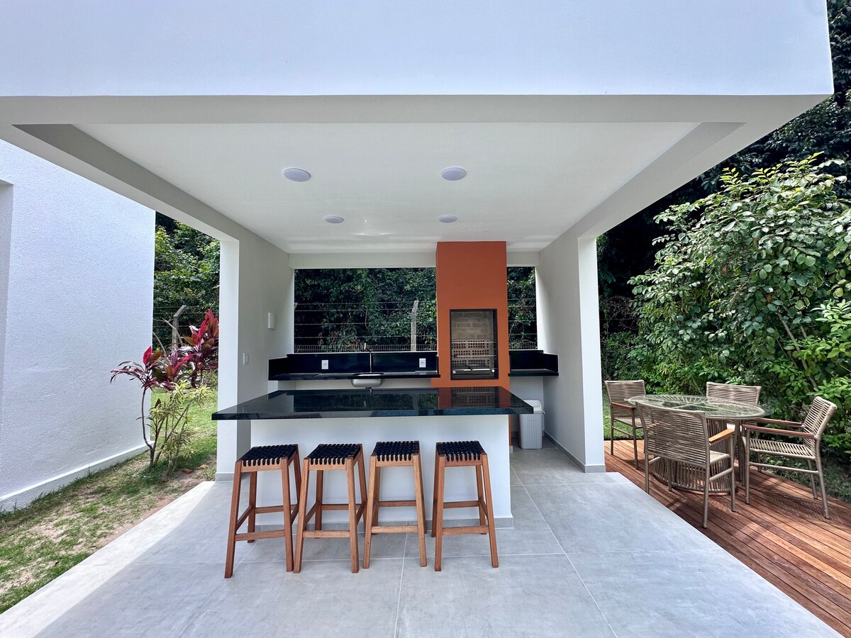 An outdoor kitchen area features a black countertop with a built-in grill, surrounded by wooden stools. To the side, a dining table with comfortable chairs is set within a serene garden, providing a space for relaxation and dining in nature.
