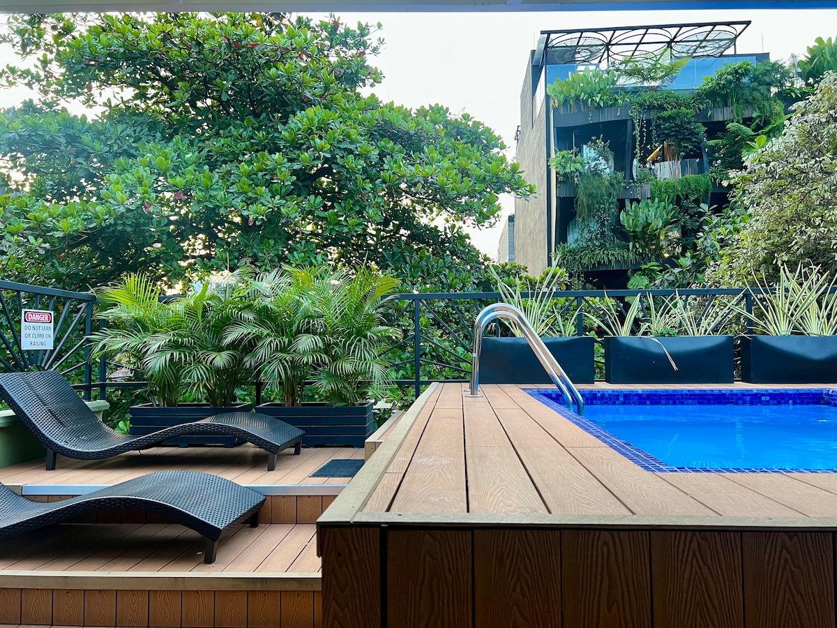 A lush rooftop area features a blue, heated pool surrounded by a wooden deck. Two black lounge chairs are positioned for relaxation, while greenery from nearby trees adds a natural backdrop. The modern building structure is visible in the background, complementing the tranquil atmosphere.