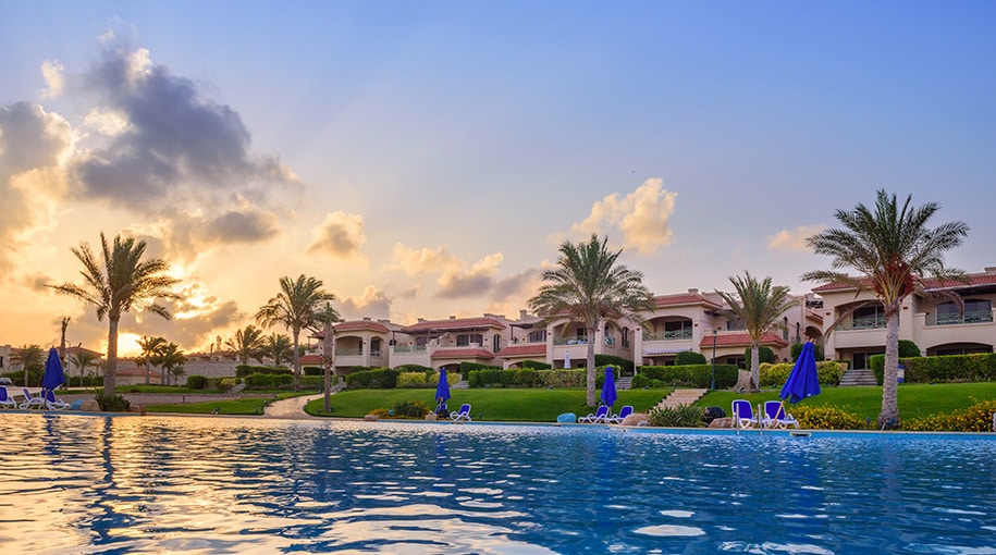 A shimmering infinity pool reflects the vibrant sunset, framed by lush greenery and palm trees. The elegant resort buildings rise in the background, each featuring spacious balconies. Comfortable lounge chairs line the pool area, inviting relaxation and enjoyment of the serene surroundings.