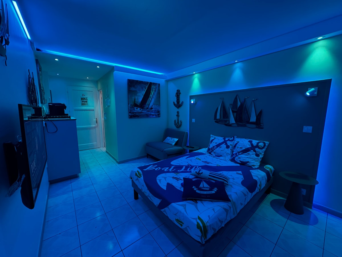 A modern studio is illuminated with soft blue LED lights, creating a calming ambiance. The room features a spacious bed adorned with patterned bedding, a cozy seating area, and nautical-themed wall art. A television is mounted on one wall, enhancing the entertainment options.