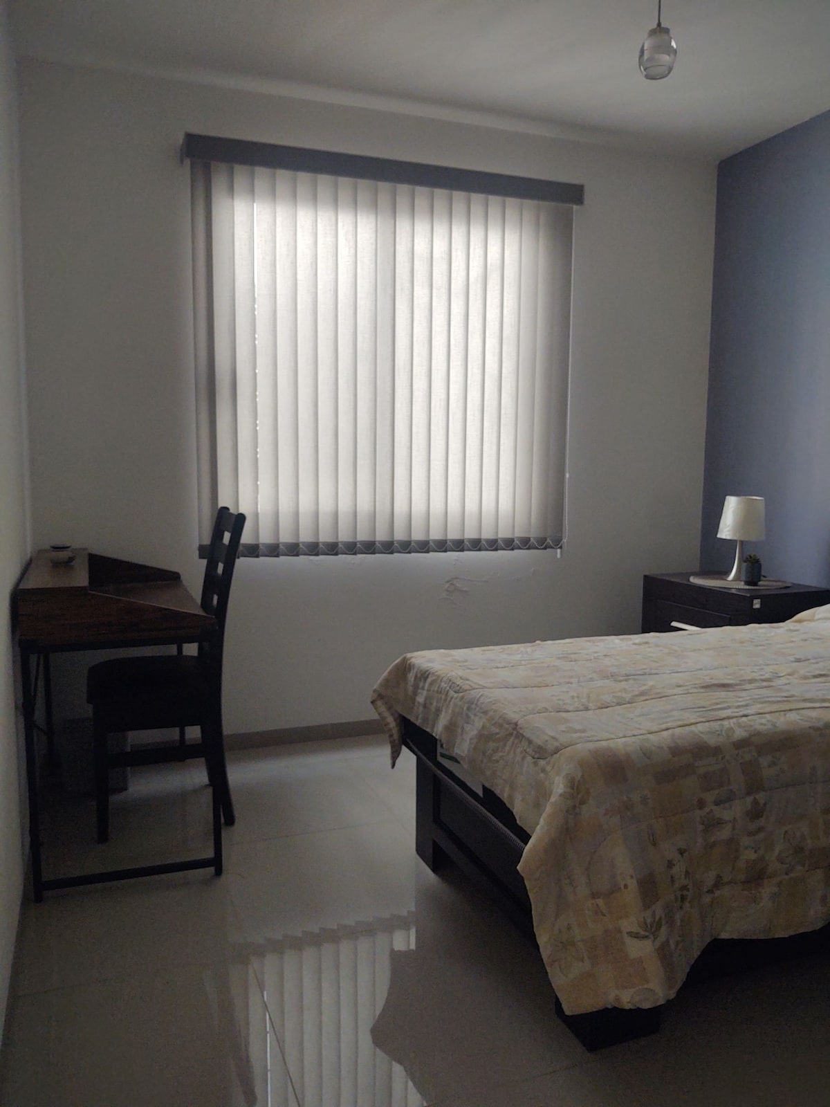 A well-lit bedroom features a spacious bed covered with a patterned bedspread. A wooden desk and chair are positioned against one wall, while a bedside table holds a lamp. Vertical blinds allow soft light to enter from the window.
