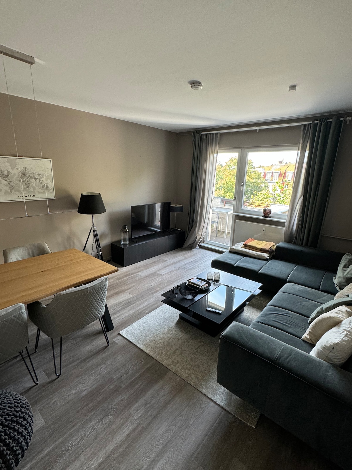 A modern living area features a dark sectional sofa, low coffee table, and a dining table with chairs. A television stands on a media console, and natural light enters through a large window, enhancing the neutral tones of the room. Decorative accents are arranged thoughtfully.