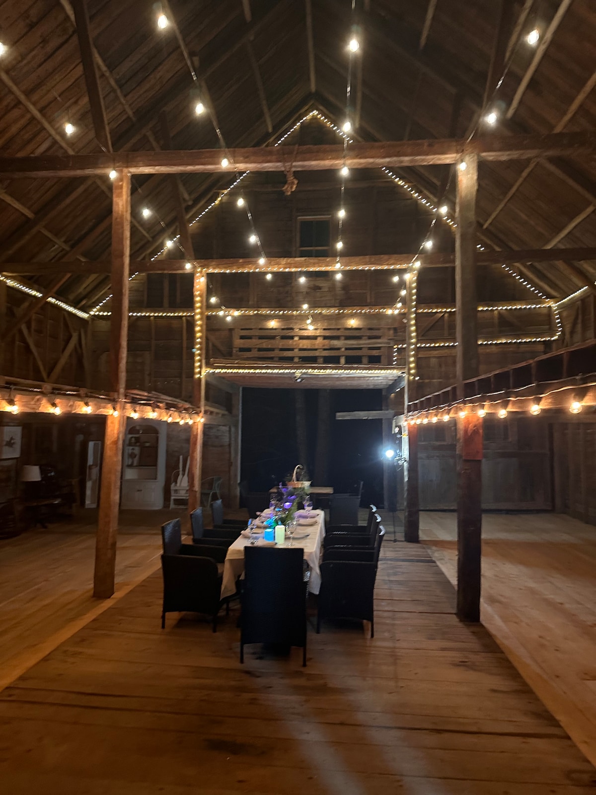 An open barn space features warm wooden beams and a polished wooden floor. Soft lighting strung across the ceiling creates a welcoming ambiance. A long dining table is set with dark chairs, ready for gatherings or celebrations.