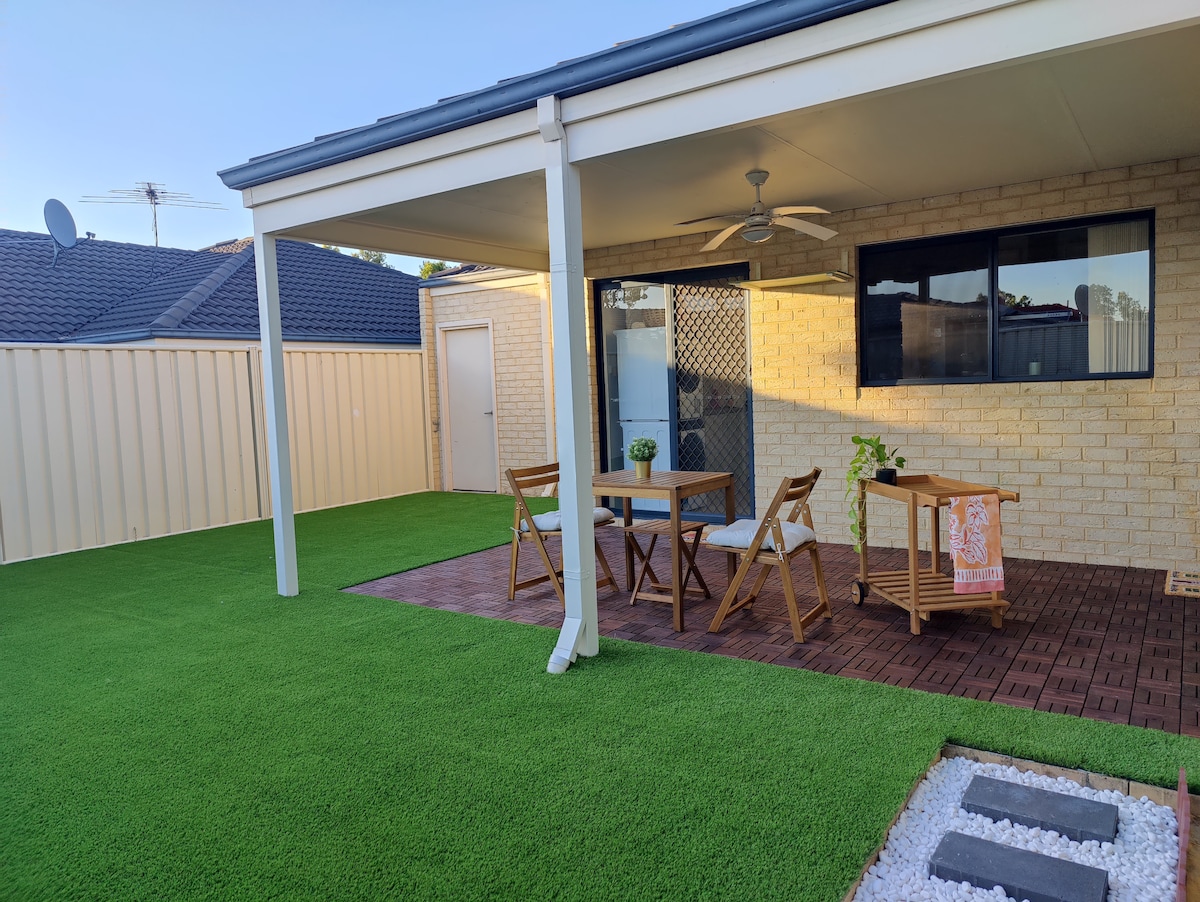 An outdoor patio area features a wooden table surrounded by four folding chairs, situated on a tiled surface. A small rolling cart holds a potted plant and a folded towel. Lush green artificial grass surrounds the space, providing a comfortable setting for relaxation.